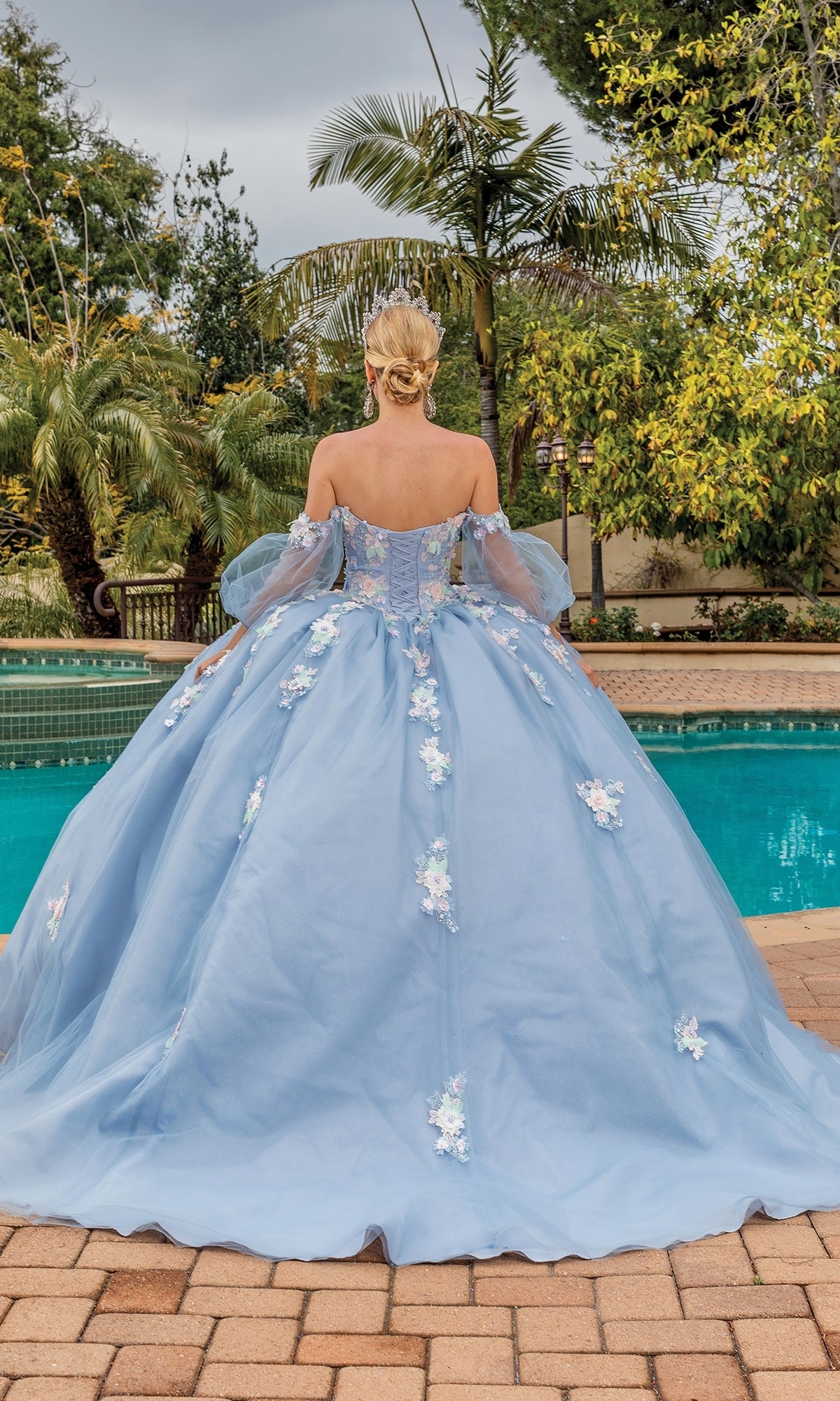 A person with blonde hair in an updo wears the Quinceanera Dress 1871 By Dancing Queen, a light blue a-line ball gown with lace floral applique, standing by a swimming pool with trees in the background.